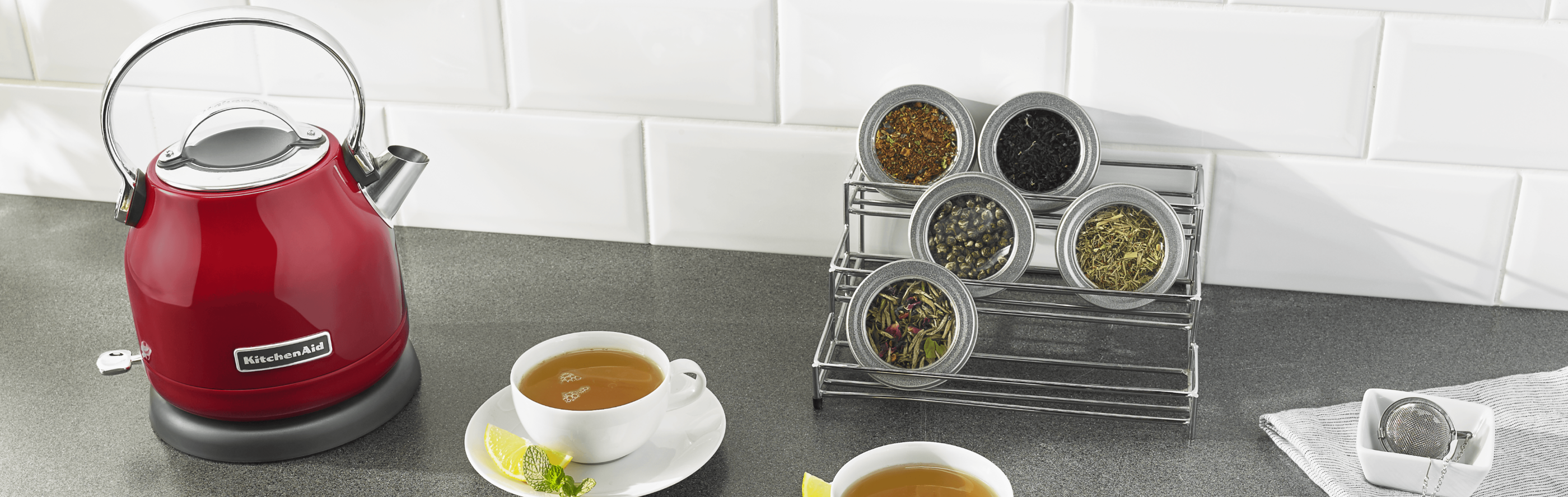 KitchenAid® electric tea kettle on countertop with cups of tea and containers of loose tea leaves.